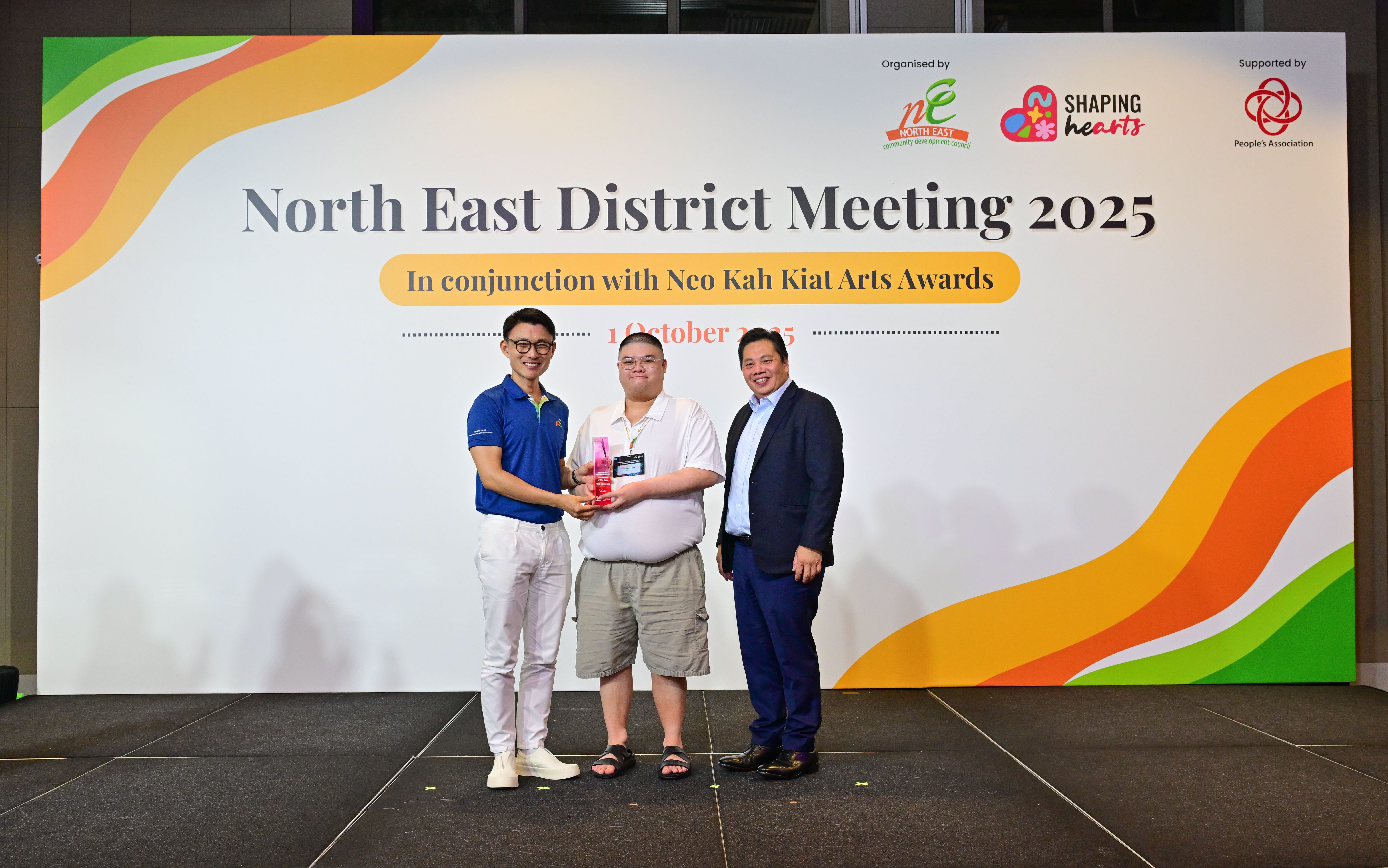 Award recipient standing on stage with the presenter, holding a acrylic plaque and posing for a group photo during the award ceremony, with a backdrop reading ‘North East District Meeting 2025 In conjunction with Neo Kah Kiat Arts Awards'